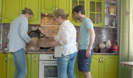 Three people frying pancake. Man standing nearby, senior woman taking pancake of a pan, young woman holding a plate. Domestic daily life, togetherness, family.の写真素材