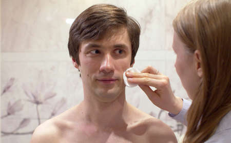 Close up portrait of young handsome man whose wife wipes his face with a cotton pad in the bright bathroom. Male face skin care. Man making funny facesの写真素材