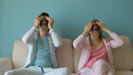 Funny couple in black facial masks putting slices of cucumbers on their eyes. Family in pajamas sitting on the couch and getting beauty treatmentsの写真素材