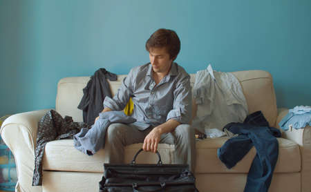 Young handsome man sitting on the sofa and packing travel bag. Couple quarrel divorceの写真素材