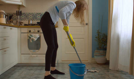 Young woman in protective yellow gloves washing the floor in the kitchen using mop. Domestic daily...