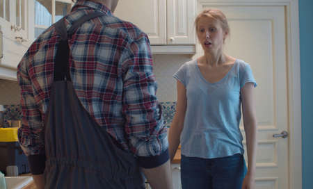 Plumber in overalls and young housewife in the kitchen. Upset woman emotionally explains something to worker. Plumbing in need of repair.の写真素材