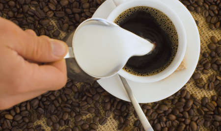 Close up roasted coffee beans and cup of fresh coffee on burlap. Hand pouring milk into coffee. Top view.の写真素材