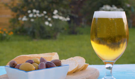 Close up lager beer in a glass. Foamy sparkling gold drink on blurry natural background outdoors. Bread and olives on the tableの写真素材
