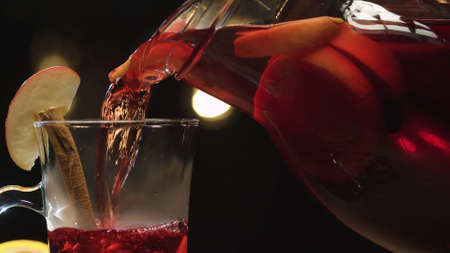 Close up hot red mulled wine pouring into a glass on black background. Slice of apple and cinnamon stick in the glass. Rising steamの写真素材