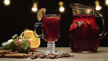 Close up hot red mulled wine in a glass with slice of apple and cinnamon stick. Jug with milled wine. Blurry lights on black backgroundの写真素材