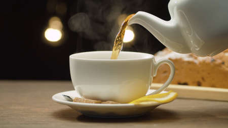 Close up hot tea pouring into a white cup. Blurry lights on black background. Steam rises above the cupの写真素材