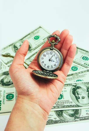 Old pocket watch hold in a hand, American dollar banknotes money on the background. Time and business concept.の写真素材