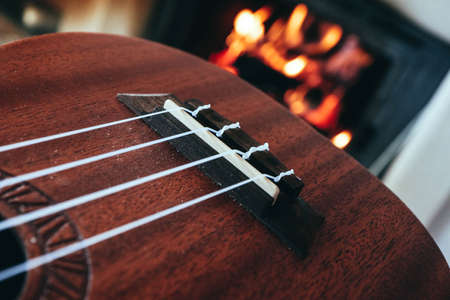 Ukulele small guitar close up stings, fireplace on the background. Musical concept, guitar fret board macro, fire in chimney, cosy romantic atmosphere.の写真素材