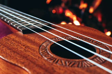 Ukulele small guitar close up stings, fireplace on the background. Musical concept, guitar fret board macro, fire in chimney, cosy romantic atmosphere.の写真素材