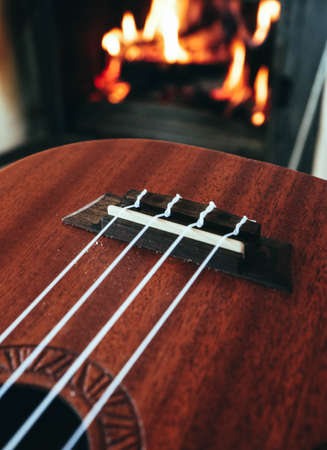 Ukulele small guitar close up stings, fireplace on the background. Musical concept, guitar fret board macro, fire in chimney, cosy romantic atmosphere.の写真素材