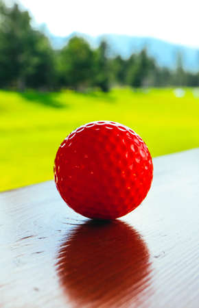 Golf red ball on a wooden surface, green field and blue sky. Macro view, luxury sport concept.の写真素材