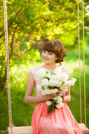Beautiful girl in a long dress with flowers in the summer on a swingの写真素材