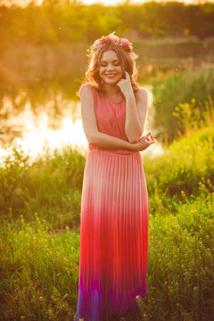 Young girl in a long dress and a flower wreath at sunset by the river in the summerの写真素材