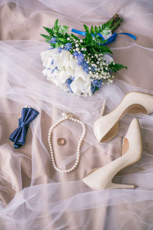 Beautiful set of women's and groom's wedding accessories. Bride's morning. Bride's bouquet of white tulips and blue flowers. The white shoes, pearl necklace and rings on white lace clothの写真素材