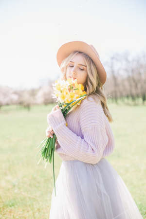A young beautiful blonde girl in a hat and a lush long skirt with bouquets of daffodils in the spring garden. Young lady holding bouquet of daffodils in springの写真素材