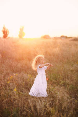 Adorable happy little girl with a curly blonde, wearing a white dress, standing in the sunny sunset field among a wild grass and flowers, smilingの写真素材