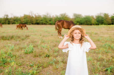 A nice little girl with a light curly hair in a white dress. Rural life in autumn. Horses and peopleの写真素材