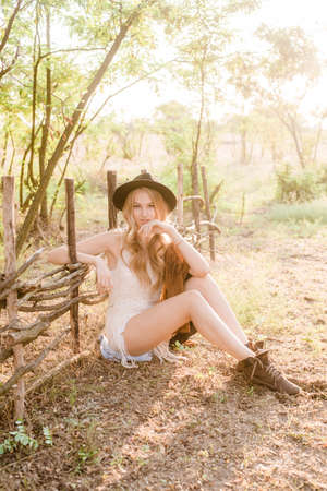 Beautiful young girl with blond hair in a suede jacket with fringed and black felt hat in the countryside on a sunny autumn day on a sunset smilingの写真素材