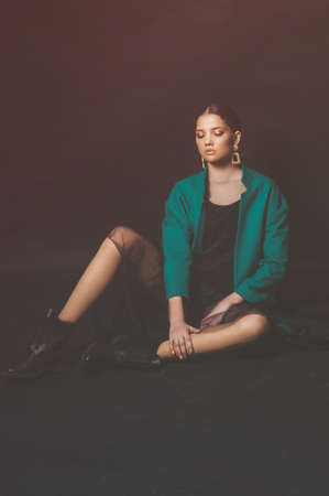 Attractive young girl model with dark hair in a trendy green jacket and jewelry on a black background in the studioの写真素材