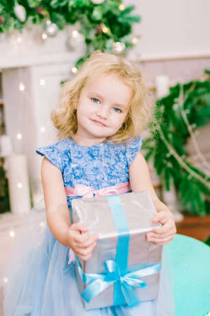 Cute girl with blond hair in a white ball gown by the Christmas tree in holiday dresses with gifts and silver confetti. Christmas moodの写真素材