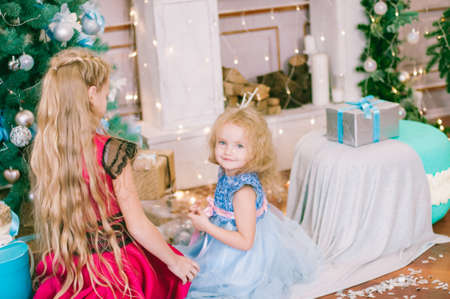 Cute girls with blond hair in a white ball gown by the Christmas tree in holiday dresses with gifts...