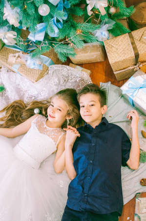 Children, brother and sister, are lying under the Christmas tree in holiday dresses with gifts and silver confetti. Christmas moodの写真素材