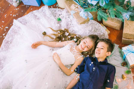 Children, brother and sister, are lying under the Christmas tree in holiday dresses with gifts and...