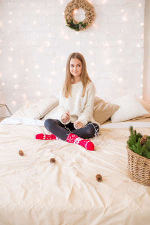 Cute teenage girl has fun at home in a light loft on the bed, decorated for Christmas with garlands and needles. Christmas moodの写真素材