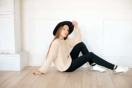 Little teenage girl with long hair in a warm knitted sweater and black felt hat in the studio on a white background. Teen fashionの写真素材