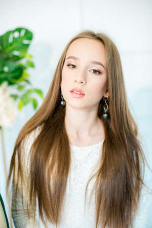 Teenager girl with long hair in a white sparkling dress with white orchids and tropical leaves in...