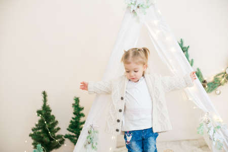 Little cute girl in a white warm knitted cardigan near a children's lodge decorated with a garland and needles, toys and tangerines. Christmas moodの写真素材