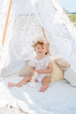 A little cute girl with red curly hair in summer, on the seashore in an airy lace bodysuit and a floral headband in a lace tent, devoried with marine details, shells and cotton. 1st BIRTHDAY.の写真素材