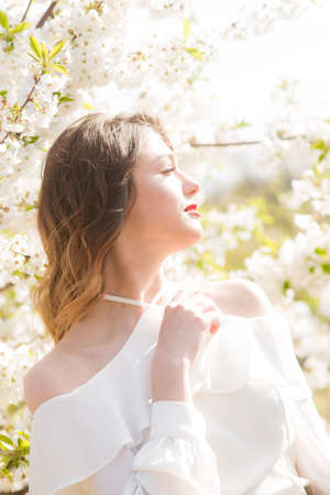 Lovely young girl in a white romantic silk blouse in spring in a blooming cherry garden in sunny weather. Spring and Fashionの写真素材