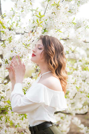 Lovely young girl in a white romantic silk blouse in spring in a blooming cherry garden in sunny...