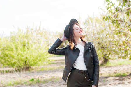 Lovely young girl in a black hat and a fashionable black leather jacket in spring in a blooming cherry garden in sunny weather with a blooming cherry branch in her hands. Spring and Fashionの写真素材