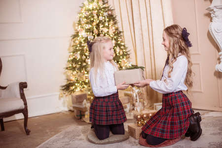 Lovely girls sisters with blonde curly hair with bans to the tols in classic school clothes by the Christmas tree in gold colors with presents in a classic interior. Christmas moodの写真素材