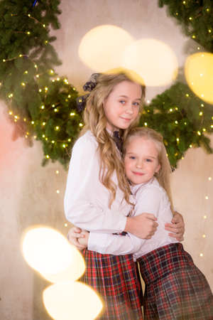 Lovely girls sisters with blonde curly hair with bans to the tols in classic school clothes next to a large Christmas wreath of pine needles on the wall in a classic interior. Christmas moodの写真素材