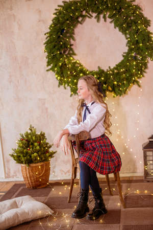 Lovely girl with blonde curly hair with bans to the tols in classic school clothes next to a large Christmas wreath of pine needles on the wall in a classic interior. Christmas moodの写真素材