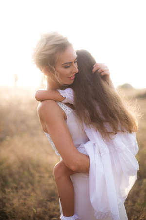 A cute little girl with long blond curly hair and her mother in a white summer dress and a straw boater hat in a field in the countryside in summer at sunset. Nature and Ecolifeの写真素材