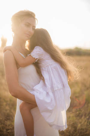 A cute little girl with long blond curly hair and her mother in a white summer dress and a straw boater hat in a field in the countryside in summer at sunset. Nature and Ecolifeの写真素材