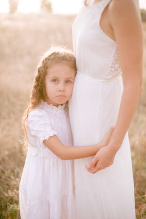 A cute little girl with long blond curly hair and her mother in a white summer dress and a straw boater hat in a field in the countryside in summer at sunset. Nature and Ecolifeの写真素材