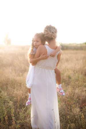 A cute little girl with long blond curly hair and her mother in a white summer dress and a straw boater hat in a field in the countryside in summer at sunset. Nature and Ecolifeの写真素材