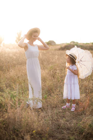 A cute little girl with long blond curly hair and her mother in a white summer dress and a straw boater hat in a field in the countryside in summer at sunset. Nature and Ecolifeの写真素材