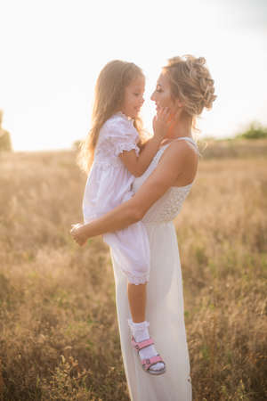 A cute little girl with long blond curly hair and her mother in a white summer dress and a straw boater hat in a field in the countryside in summer at sunset. Nature and Ecolifeの写真素材