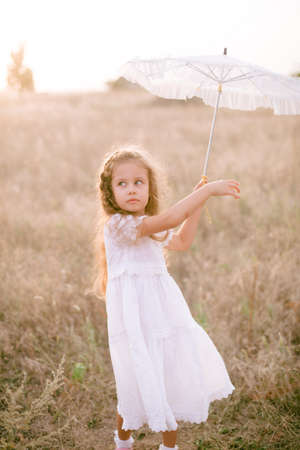 A cute little girl with long blond curly hair in a white summer dress and a straw boater hat with a white lace umbrella in a field in the countryside in summer at sunset. nature anの写真素材