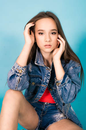 A cute teenage girl with long hair in trendy denim youth clothes and a red T-shirt on a blue...