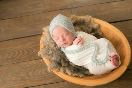 A cute baby sleeps wrapped in an airy milk-colored blanket in a wooden basket and blue knitted cap at home.の写真素材