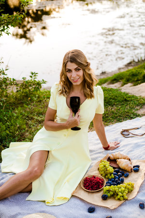 Young attractive girl with dark hair in a yellow summer dress on a picnic in nature by the river in summer at sunset. Healthy life.の写真素材