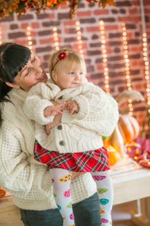 A young mother and little daughter in white warm knitted sweaters in an interior decorated with blankets and pillows as well as pumpkins, autumn leaves and apples. autumn mood. halの写真素材